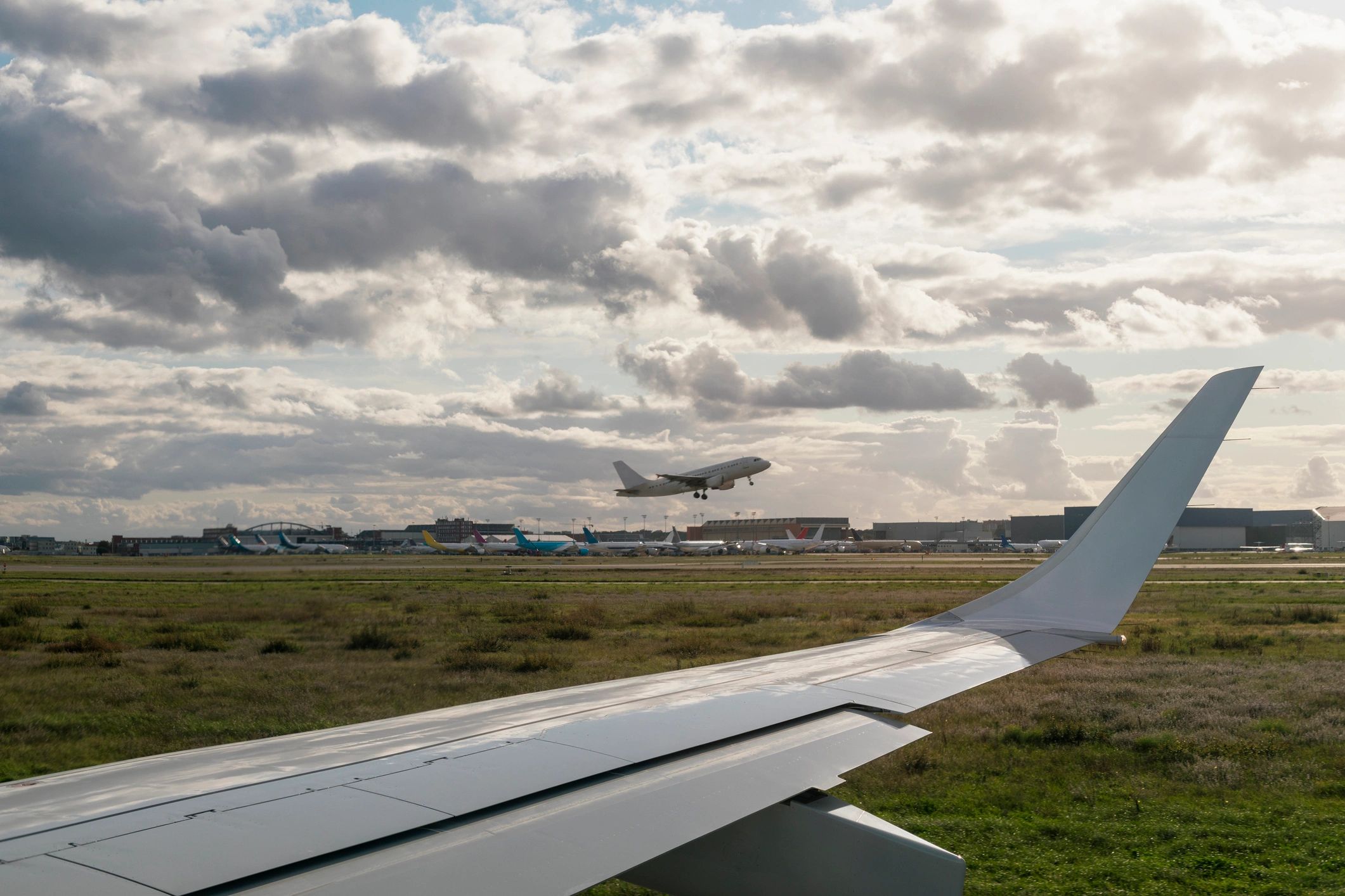 Airplane wing on runway with another aircraft taking off